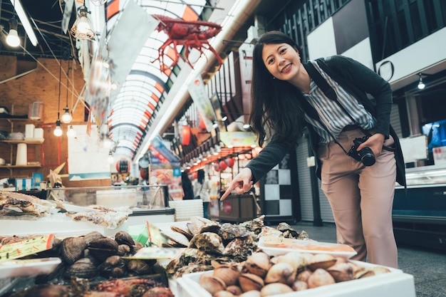 写真 大阪の黒門市場で新鮮な魚介類を売っている新鮮な魚介類を売っている屋台の食べ物を売っている屋台を示す明るい笑顔のポインティングカメラを持つアジアの若い女性のビンテージ写真。