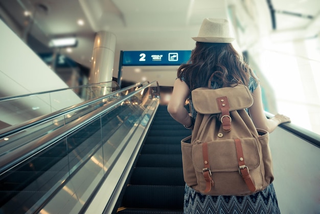 빈티지 레트로 필름 컬러 사진입니다. 배낭을 메고 공항에 도착했을 때 게이트로 가는 전기 에스컬레이터에 서 있는 여성 여행자의 뒷모습.
