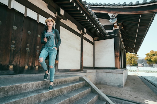 vintage stijl van volle lengte lachende hipster vrouw reiziger met camera traplopen tijdens toeristische tour in osaka japan. positieve vrouwelijke fotograaf sightseeing prachtig uitzicht op shitennoji.