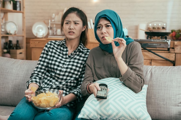 Vintage style of two asian women roommates with bowl of potato
chips watch horror film on sofa in home kitchen. young girl friends
enjoy day free time together watching thriller film on
television.