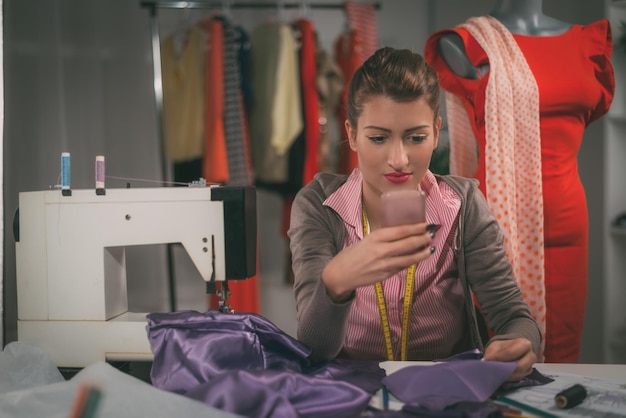 Vintage toned photo of a young woman designer sits in front of the sewing machine and using mobile phoning.