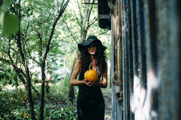 Vintage vrouw als heks poseren met pompoen in de handen tegen verlaten plek aan de vooravond van Halloween