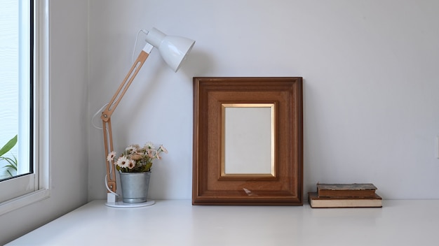 Vintage workspace with empty frame, pot plant, lamp and old\
book on white table.