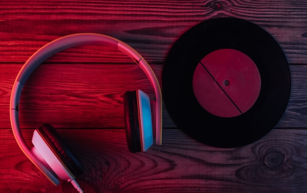 Vinyl record, stereo headphones on wooden surface. Neon red and blue light