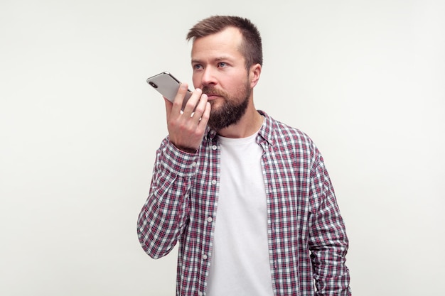 Virtual assistant. Portrait of serious bearded man in casual plaid shirt talking to mobile phone using voice application, audio message or recorder. indoor studio shot isolated on white background