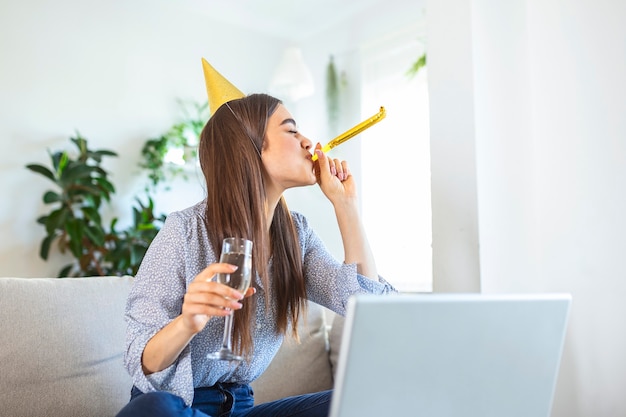 Virtual celebration. happy woman in party hat celebrating\
birthday online at quarantine or self-isolation, using laptop for\
video call with friends and family, holding champagne