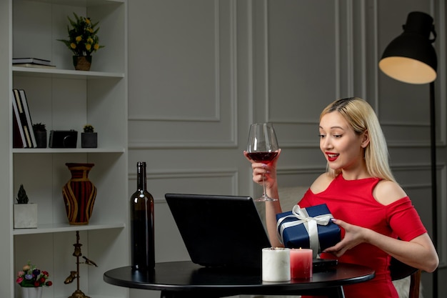 Virtual love cute blonde girl in red dress on distance date with wine holding wine and gift box