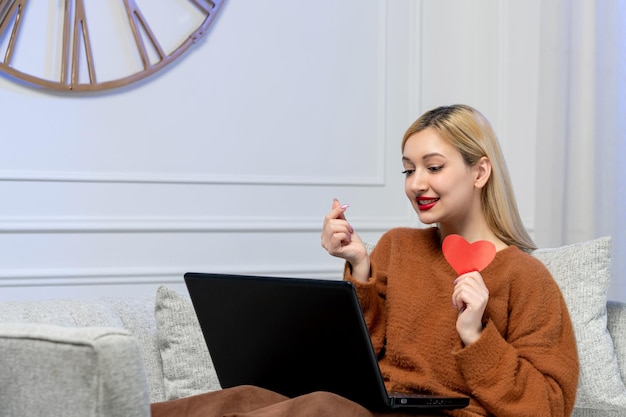 Virtual love cute young blonde girl in cozy sweater on computer distance date holding paper heart