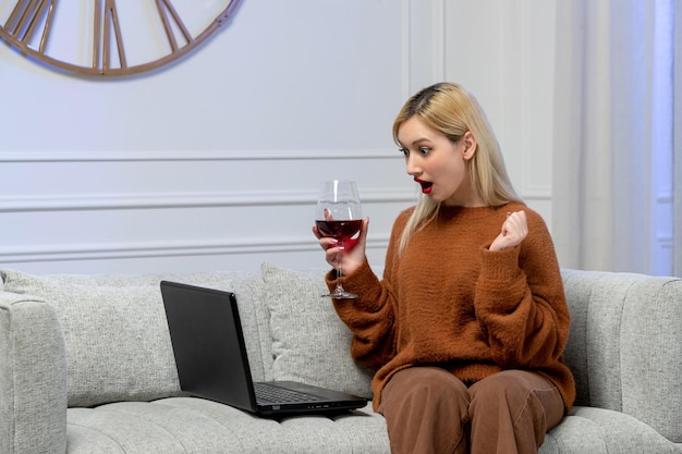 Virtual love cute young blonde girl in cozy sweater on distance computer date holding fist and wine