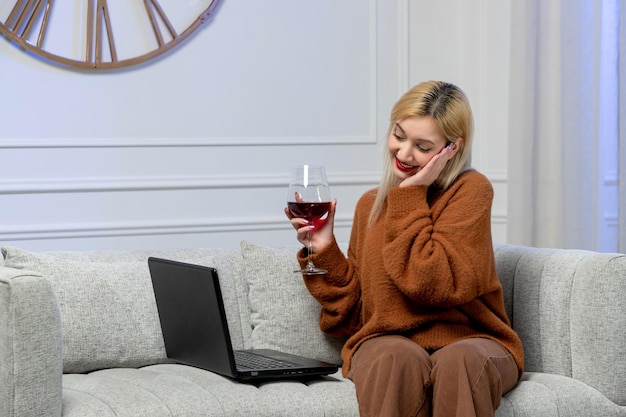 Virtual love cute young blonde girl in cozy sweater on distance computer date holding head and wine