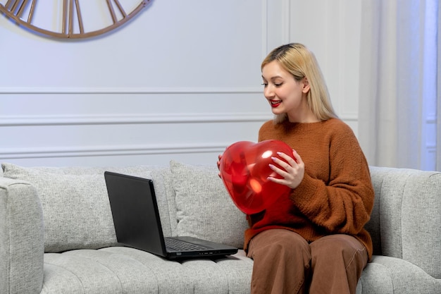 Virtual love cute young girl in cozy sweater on distance computer date with red heart balloon