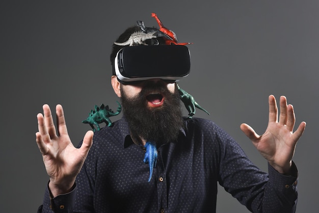 Virtual reality, 3d technology, entertainment, gaming, travel concept - bearded man with virtual reality goggles traveling in jurassic park. Era of dinosaurs. Jurassic period. Man with toy dinosaurs.