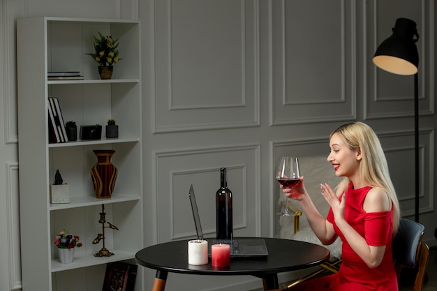 Virtueel liefdes schattig blond meisje in rode jurk op afstandsdatum met kaarsen met wijn