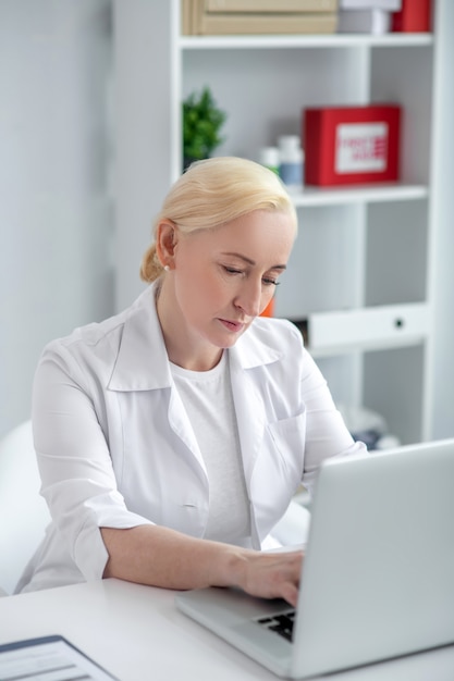 Virtuele dokter. Blonde artsenzitting op middelbare leeftijd bij haar bureau en het spreken aan patiënt op Internet