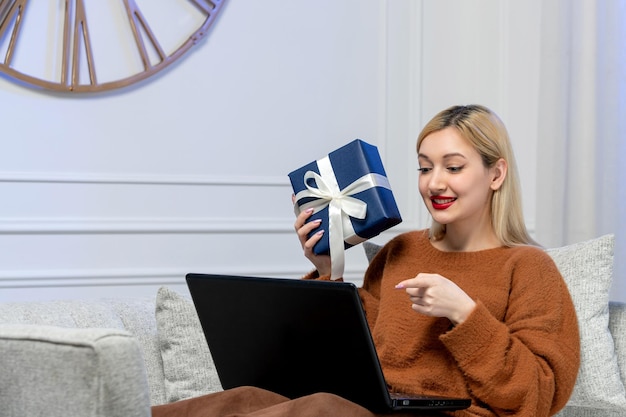 Virtuele liefde schattig jong blond meisje in een gezellige trui op computer afstandsdatum met blauwe geschenkdoos