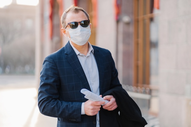 Virus, pandemic, work concept. Male manager wears formal suit and sunglasses, protective medical mask to prevent coronavirus, holds papers in hands, poses outdoor near office, waits for colleague