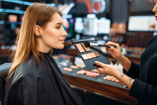 Visagiste and lady chooses cosmetic in makeup shop. Female client in beauty salon, make-up procedure