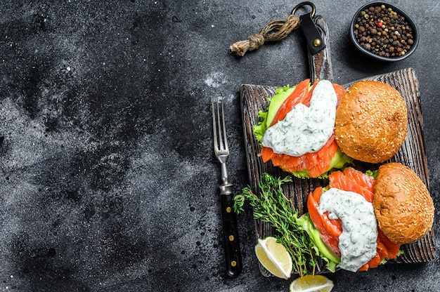 Visburger met gezouten zalm, avocado, mosterdsaus, komkommer en ijsbergsalade. Zwarte achtergrond. bovenaanzicht. Kopieer ruimte.