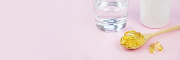 Visgelatine softgels omega3 in houten lepel, een glas water en een open witte pot met capsules op een roze achtergrond