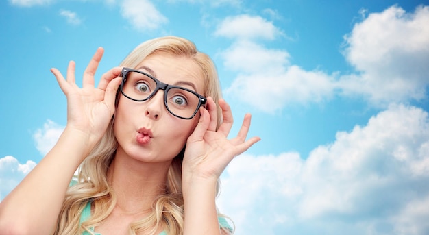 vision, education and people concept - happy young woman or teenage girl glasses making funny fish face over blue sky and clouds background