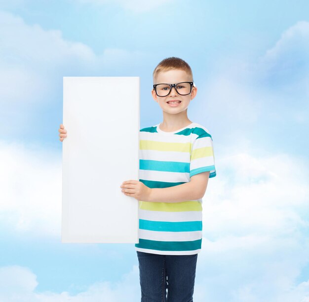 vision, health, advertisement, childhood and people concept - smiling little boy in eyeglasses with white blank board over blue cloudy sky background