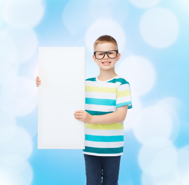 vision, health, advertisement and people concept - smiling little boy in eyeglasses with white blank board over blue background