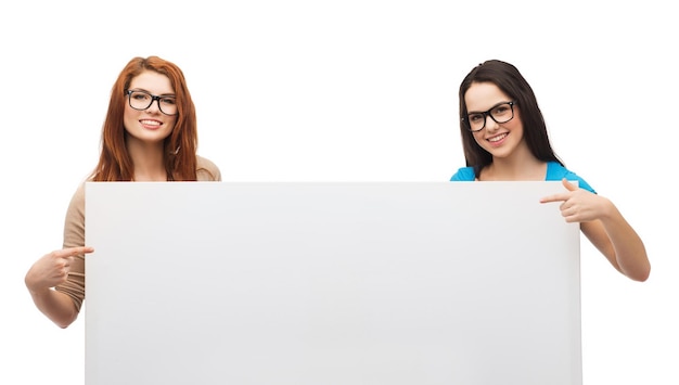 vision, health, advertisement and people concept - two smiling girls wearing eyeglasses pointing fingers to white blank board