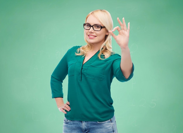 Vision, optics, education, gesture and people concept - smiling young woman with eyeglasses showing ok over green school chalk board background