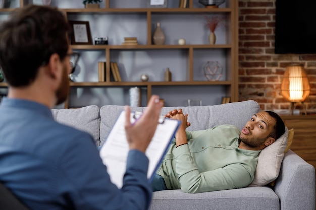 ハンサムな男がソファに横たわって話しているアフリカ系アメリカ人の心理学者への訪問