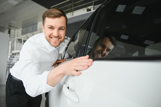 Foto in visita alla concessionaria di automobili bell'uomo barbuto sta accarezzando la sua nuova auto e sorridendo