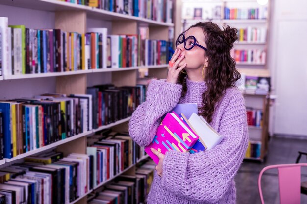 Visiting public library . Excited good-looking girl spending time between bookshelves while choosing reading for education