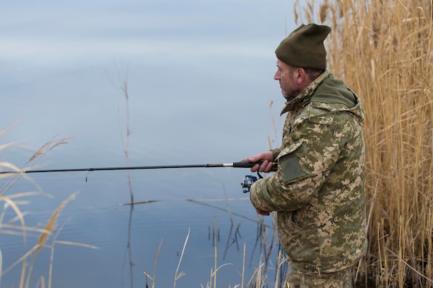 Vissen in het riet om te spinnen op rustig water Een man in camouflagekleding is bij koud weer aan het vissen op de oever van de rivier