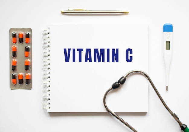 VITAMIN C is written in a notebook on a white table next to pills and a stethoscope. Medical concept