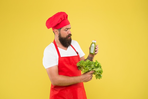 ビタミンスムージードリンクヒップスタースムージー飲料ボトル入りの緑フレッシュジュース絞りスムージー男のひげを生やしたシェフがレタスとスムージーボトルを保持黄色の背景ベジタリアン栄養