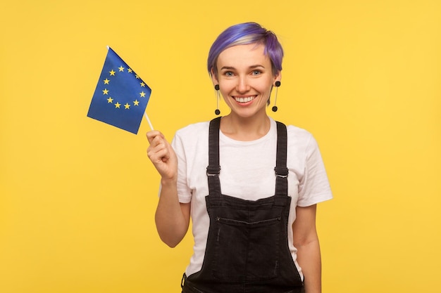 Foto vlag van de europese unie. portret van een vrolijk hipstermeisje met violet kort haar in een denimoverall die de eu-vlag vasthoudt en naar de camera kijkt met een brede glimlach. geïsoleerd op gele achtergrond, studio-opname