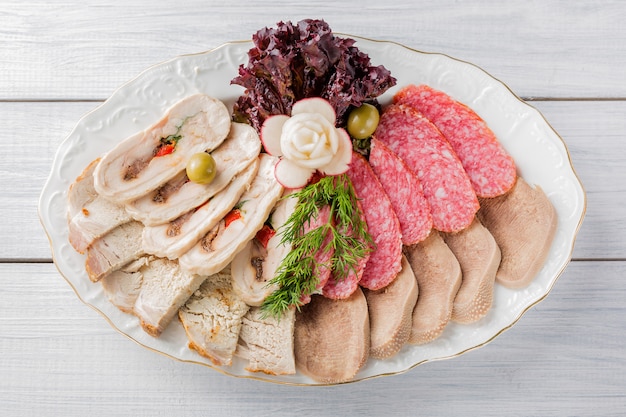 Vleesbord met heerlijke stukjes gesneden ham, worst, olijven, rundvleestong, kruiden en vlees met radijs op wit bord en houten tafel