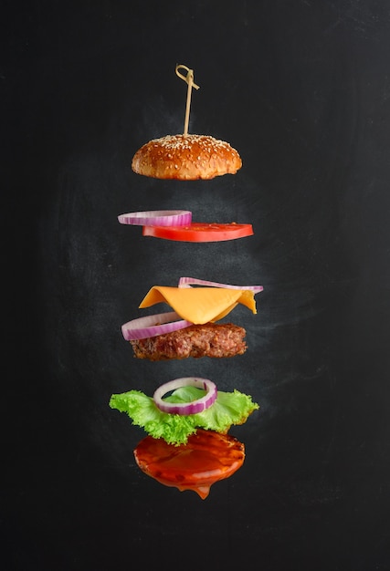 Vliegende ingrediënten van een klassiek cheeseburger sesambroodje, uienringen, plakjes tomaat en een sappige barbecuekotelet. lagen fastfood op zwarte krijtbord achtergrond