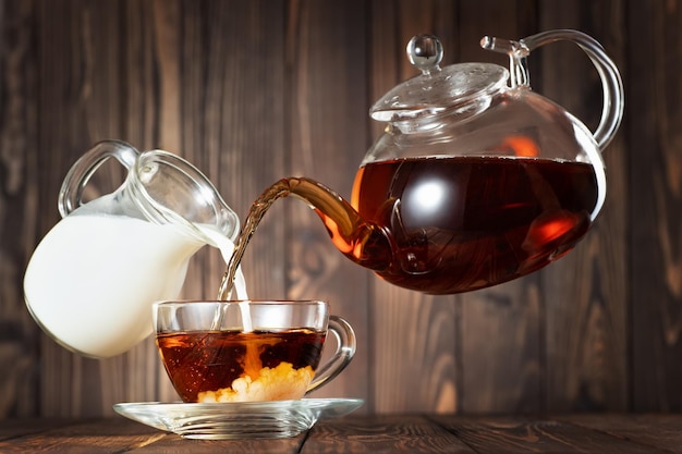 vliegende theepot en kruik die thee met melk giet in de beker op houten tafel