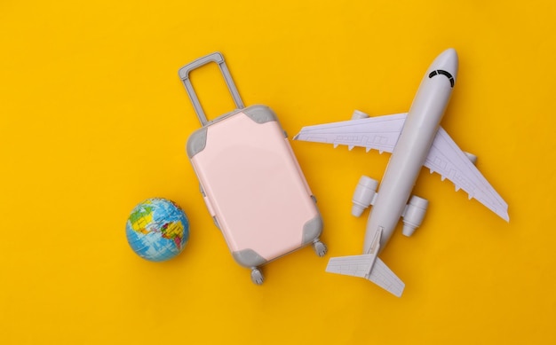 Vliegreizen plat leggen. Mini plastic reiskoffer, vliegtuig en globe op gele achtergrond. Bovenaanzicht