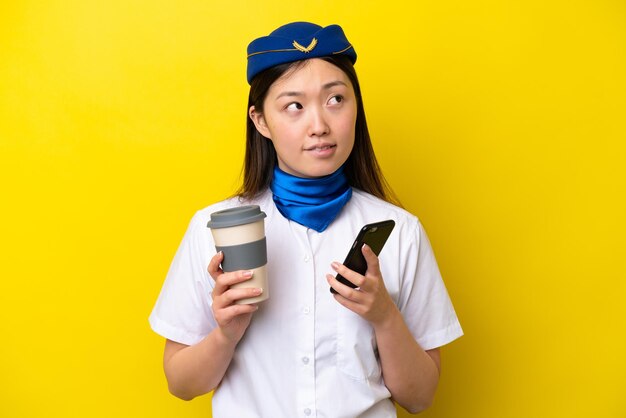 Vliegtuig Chinese vrouw stewardess geïsoleerd op gele achtergrond met koffie om mee te nemen en een mobiel terwijl ze iets denkt