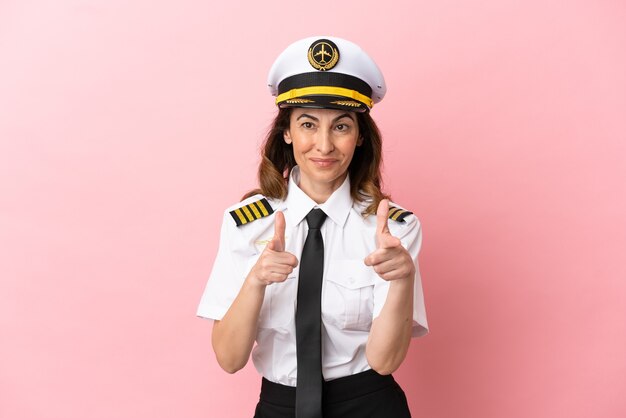 Vliegtuig piloot vrouw van middelbare leeftijd geïsoleerd op roze achtergrond naar voren wijzend en glimlachend