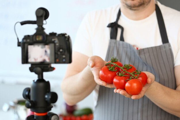 Vlogger-chef-kok die rijpe tomateningrediënt toont. man in schort opname van grote rode groente op camcorder voor culinaire vlog.