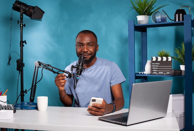 Vlogger holding smartphone looking at camera with hand on\
microphone sitting at podcast recording desk. influencer talking\
into professional mic after reading messages from fans on\
mobile.