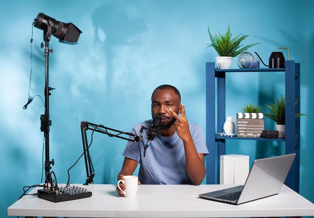 Vlogger showing peace sign sitting while holding cup at desk in recording studio with professional microphone and video light. Content creator doing hand gesture in front of audio podcast setup.