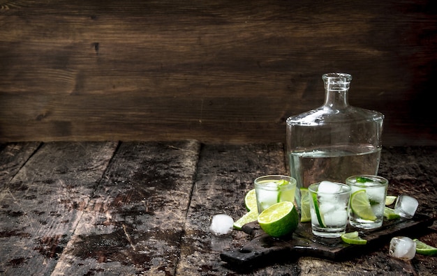 Vodka shots with lime and ice on the board on a wooden table.