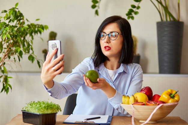 Voedingsdeskundige, diëtist die op een smartphone haar vlog opneemt over gezond eten, gezondheidszorg en dieetconcept. Vrouwelijke voedingsdeskundige met fruit die thuis bij haar des werkt.