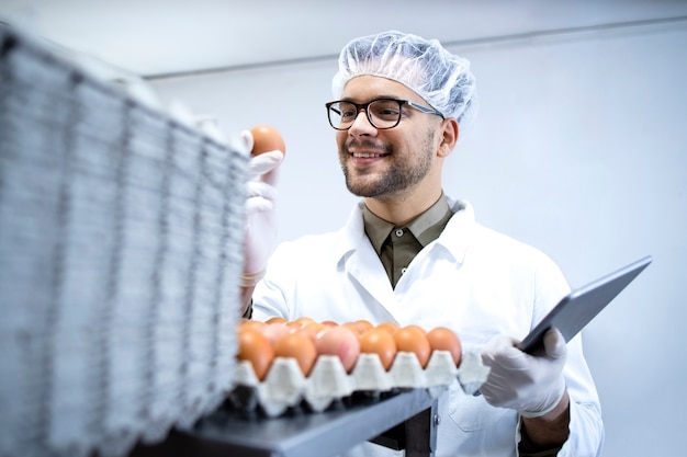 Voedsel fabrieksarbeider in witte jas haarnetje en hygiënische handschoenen eieren kwaliteit controleren bij de voedselverwerkende fabriek op tabletcomputer.