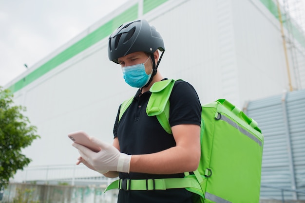 Voedsel levering concept. De bezorger van eten gebruikt een smartphone om klanten sneller te bereiken. Courier heeft een koelkast in een groene rugzak. Hij draagt een medisch masker en handschoenen