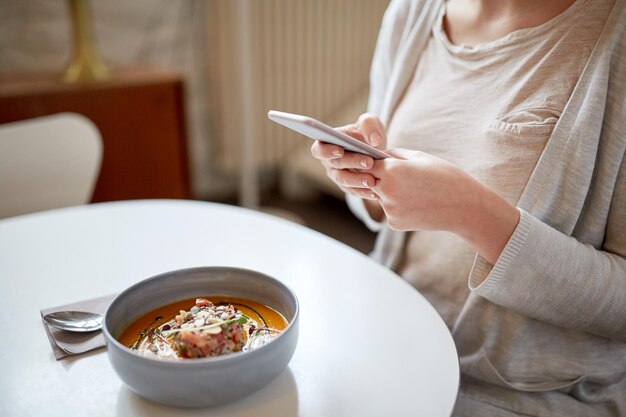 voedsel, nieuwe noordse keuken, technologie, eten en mensenconcept - vrouw zittend aan cafétafel met smartphone en kom groentepompoen-gembersoep met geitenkaas en tomatensalade met yoghurt