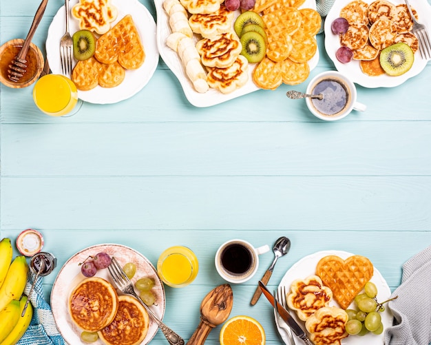 Voedseloppervlak met gezond ontbijt met verse warme wafelsharten, pannenkoekenbloemen met bessenjam en fruit op turkooise tafel, bovenaanzicht, plat leggen, kopie ruimte